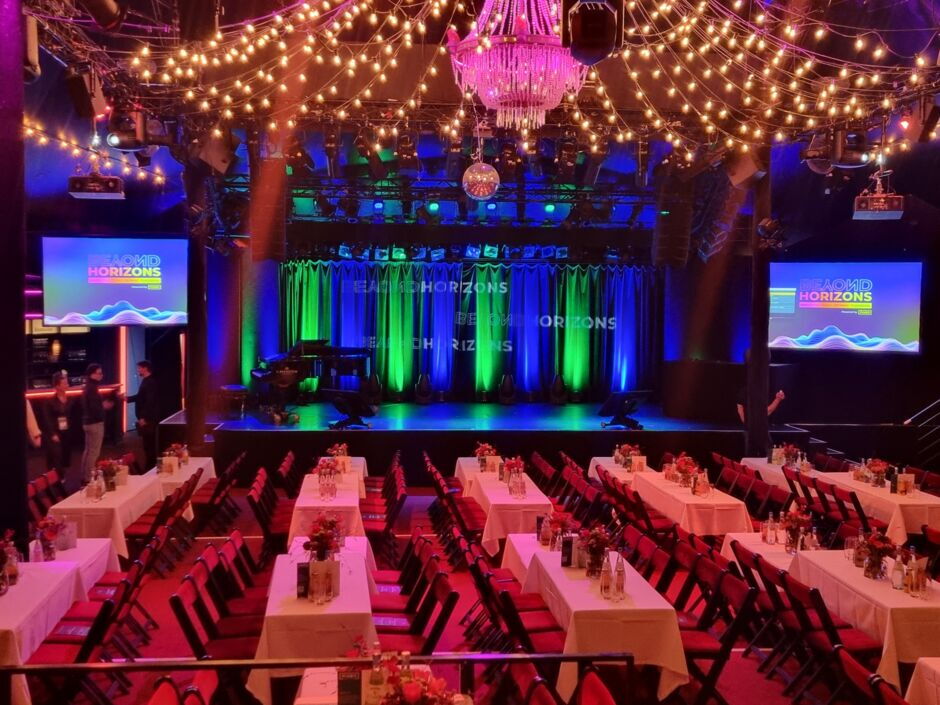 Veranstaltungssaal mit Bühne, Tischen mit weißen Tischdecken und Stühlen, beleuchtet von vielen Lichterketten und einem Kronleuchter