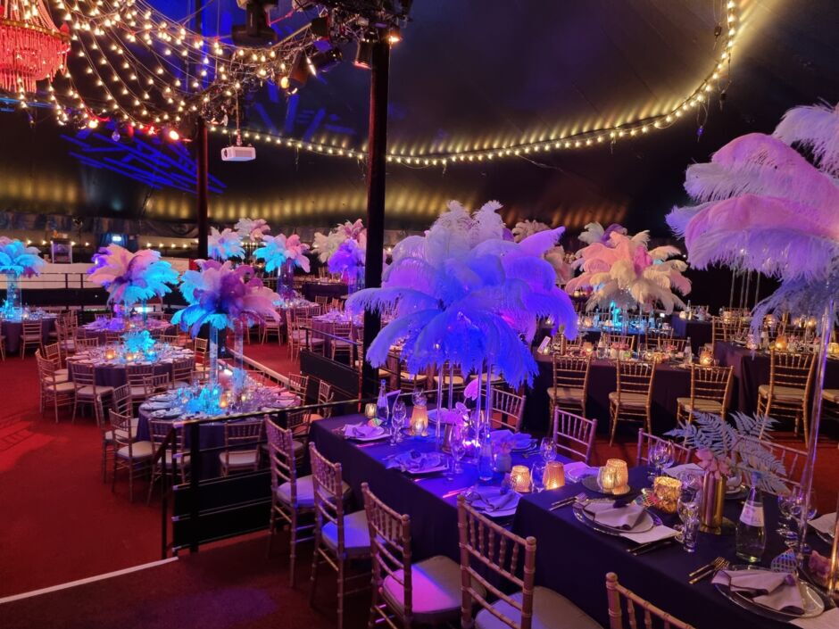 Festlich dekorierter Veranstaltungsraum mit Tischen, Stühlen und großen Federarrangements als Tischdekoration unter einem Zelt mit Lichterketten.