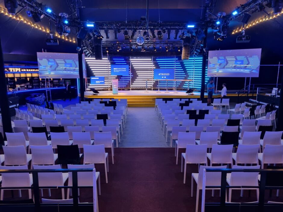 Blick auf einen Veranstaltungsraum mit Reihen von weißen Stühlen vor einer Bühne mit großen Bildschirmen und Beleuchtung.