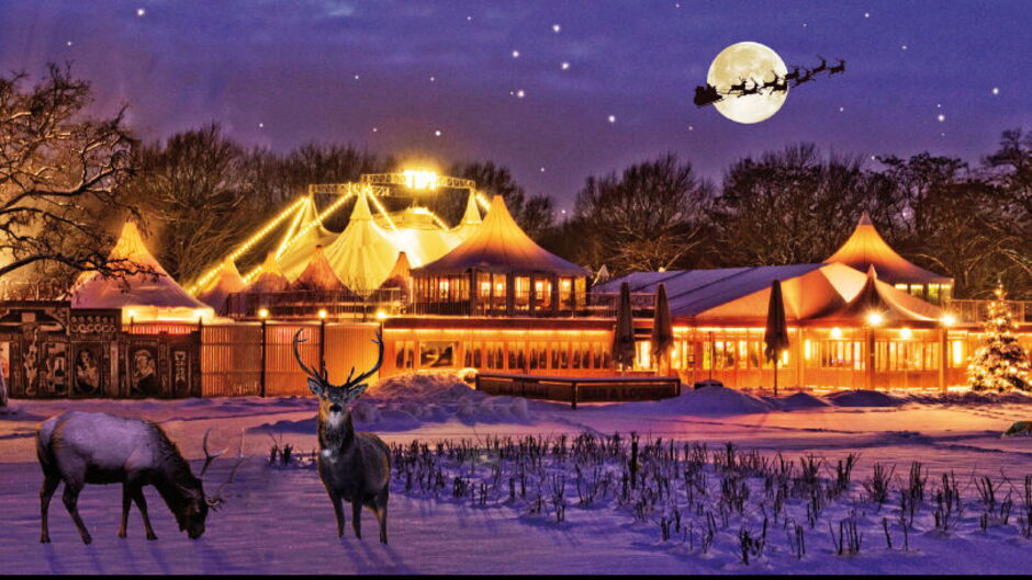 Weihnachtlich beleuchtetes Theaterzelt mit spitzen Dächern in verschneiter Landschaft, zwei Hirsche im Vordergrund, Vollmond mit Santa und Rentierschlitten am Himmel.