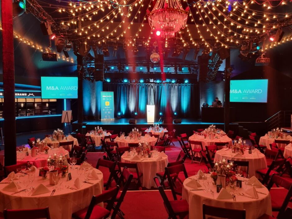 Veranstaltungsraum mit runden, gedeckten Tischen und Stühlen vor einer Bühne mit zwei Bildschirmen, die 'M&A AWARD' anzeigen, und einer Decke mit Lichterketten und einem Kronleuchter