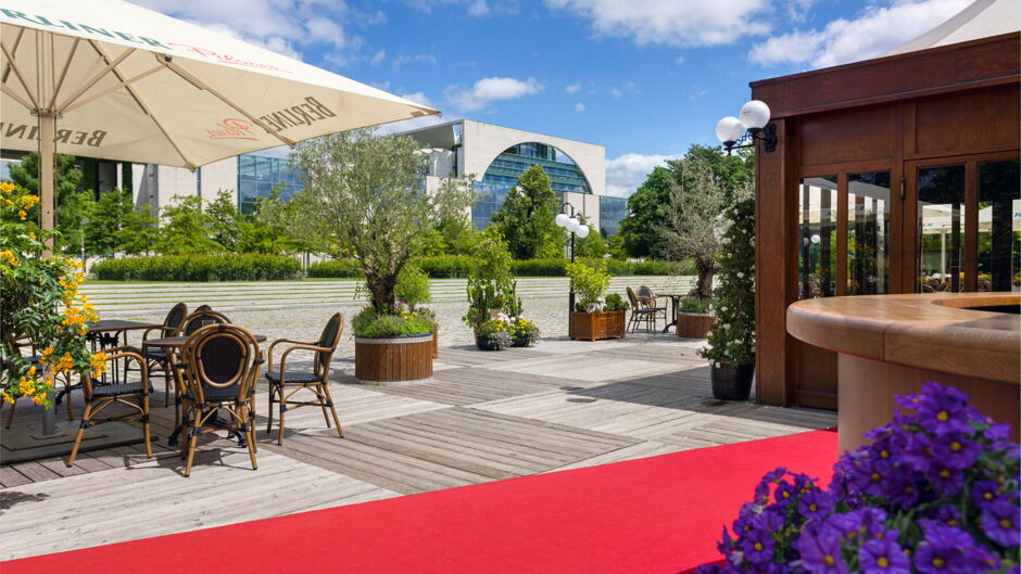 Outdoor seating area of TIPI AM KANZLERAMT beer garden with wooden deck, tables, chairs, umbrella, and view of the modern Kanzleramt building.