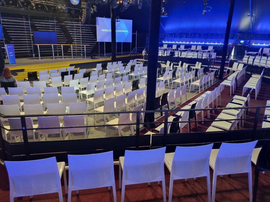 Leerer Veranstaltungsraum mit Reihen weißer Stühle vor einer Bühne und zwei Bildschirmen mit der Aufschrift 'HERZLICH WILLKOMMEN'