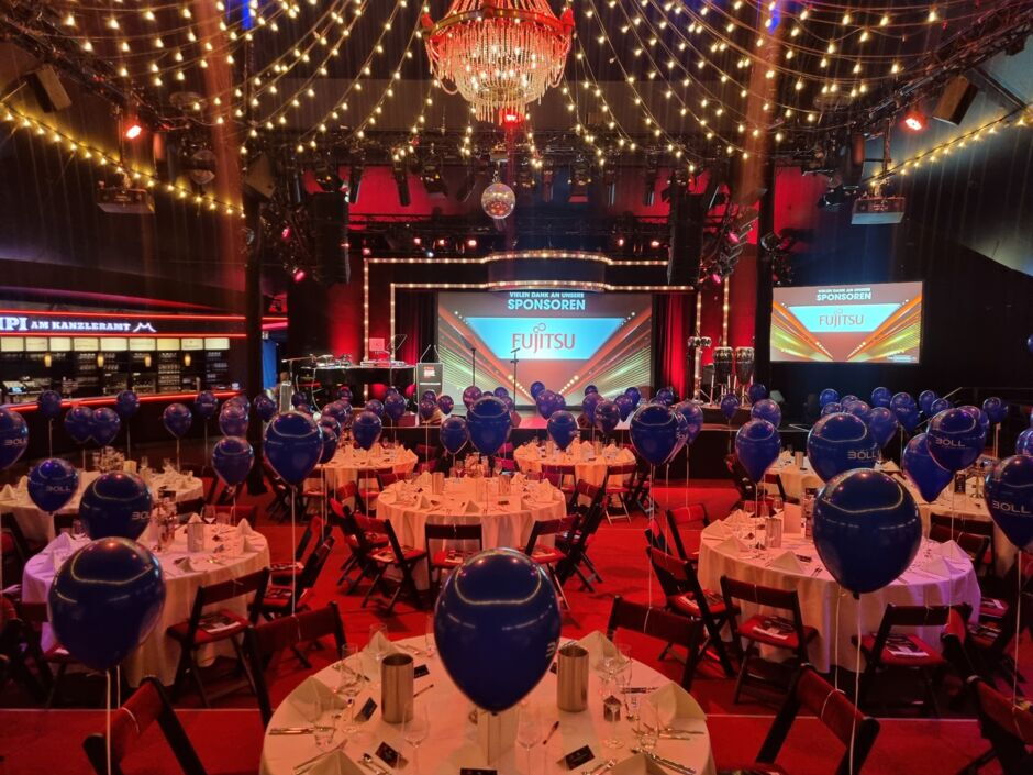 Festlich dekorierter Veranstaltungsraum mit runden Tischen, blauen Luftballons und einer Bühne mit zwei großen Bildschirmen 