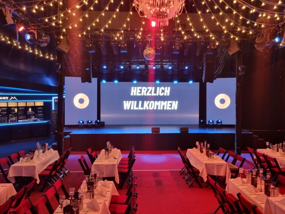 Blick auf einen festlich dekorierten Veranstaltungsraum mit Tischen, Stühlen und einer Bühne, auf der 'HERZLICH WILLKOMMEN' auf einer großen Leinwand steht.