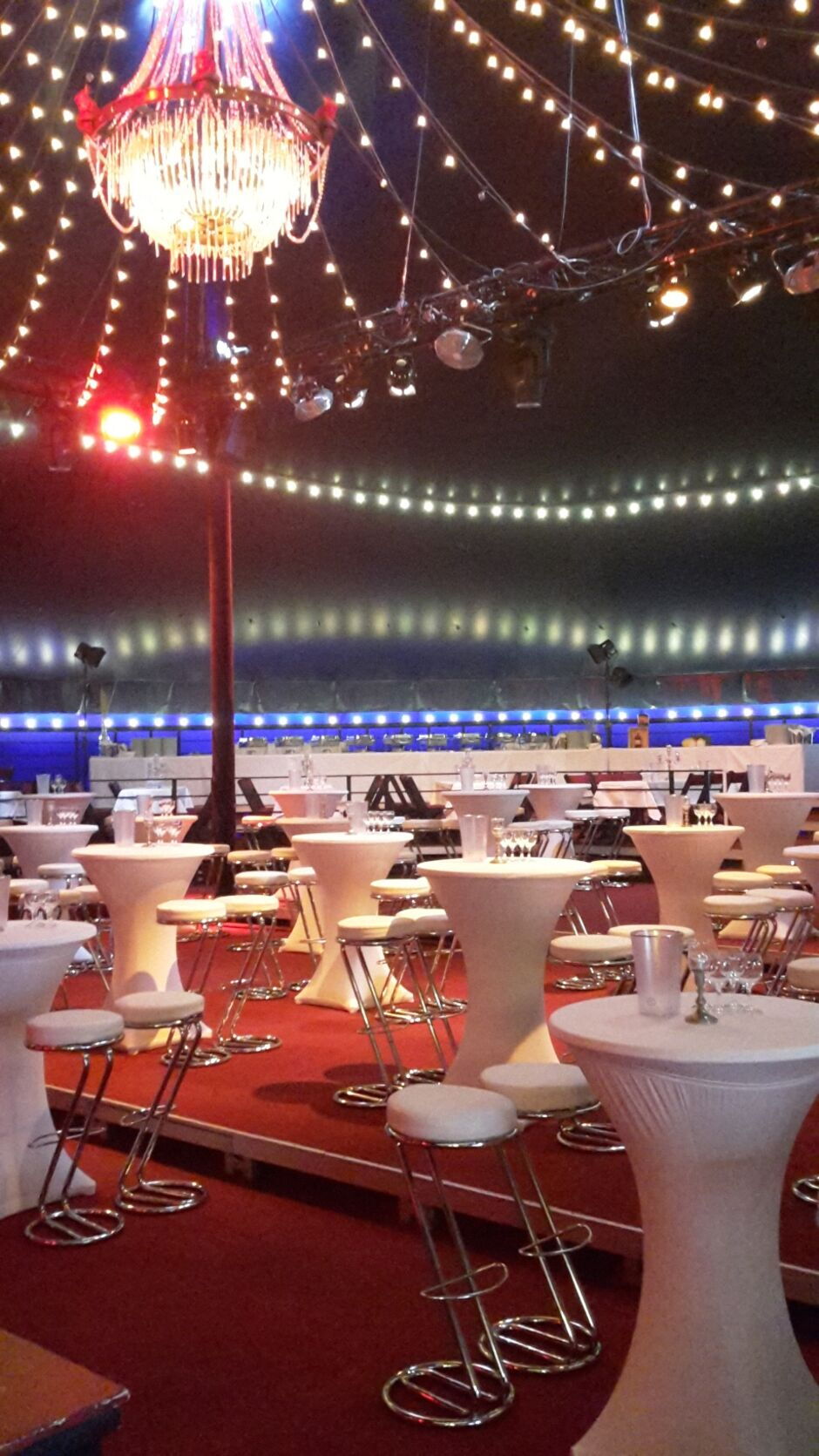 Festlich dekorierter Innenraum mit Stehtischen, Hockern und einer großen Kronleuchterbeleuchtung unter einer Zeltdecke mit Lichterketten