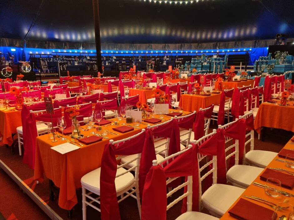Mehrere lange Tische mit orangen Tischdecken und weißen Stühlen mit roten Hussen in einem großen, rund wirkenden Raum mit blauer Beleuchtung