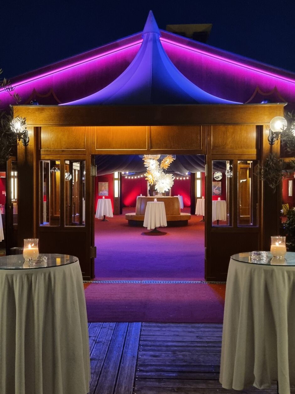 Blick durch einen offenen Holzeingang auf einen festlich beleuchteten Innenraum mit Stehtischen und Dekoration unter einem Zelt mit lila Beleuchtung