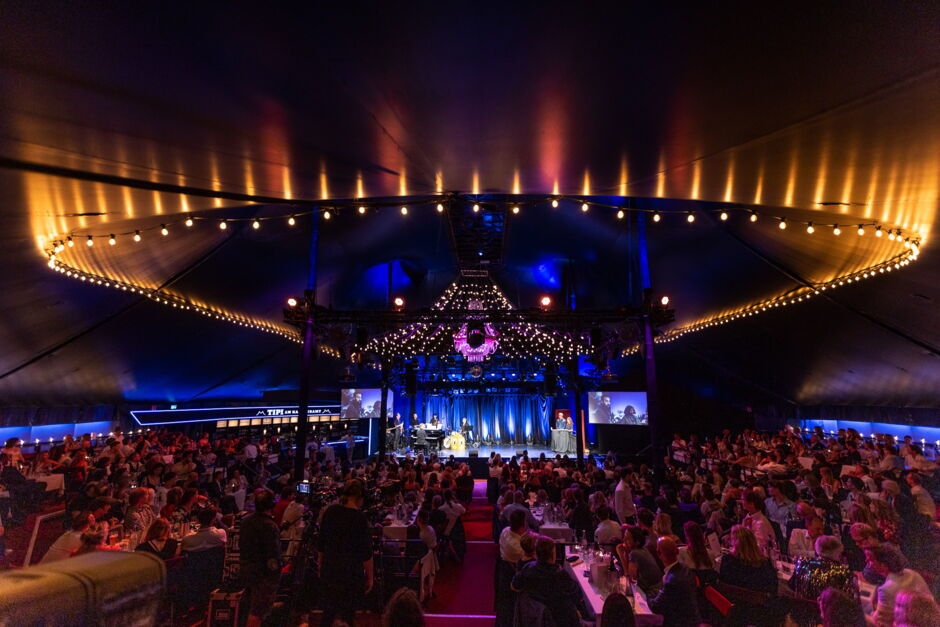 Großes Zirkuszelt mit Bühne, umgeben von Publikum an Tischen, beleuchtet mit vielen Lichtern am Zeltdach und auf der Bühne